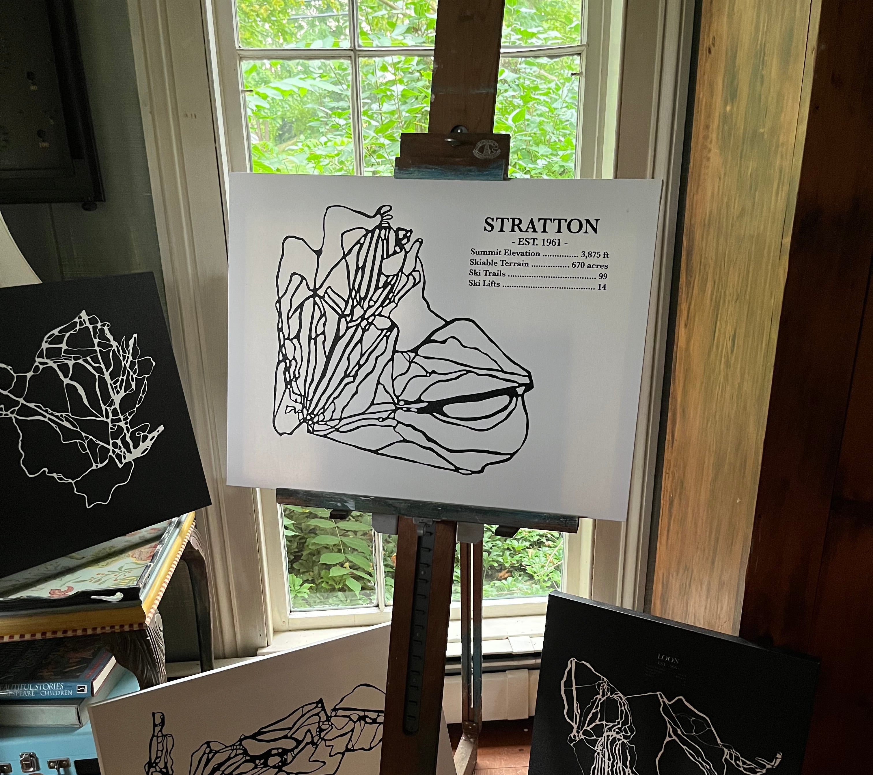 An easel in a real indoor room displays art prints and sketch cards. The central white print on the easel shows a black line drawing of a shell with the name STRATTON and date 1950.